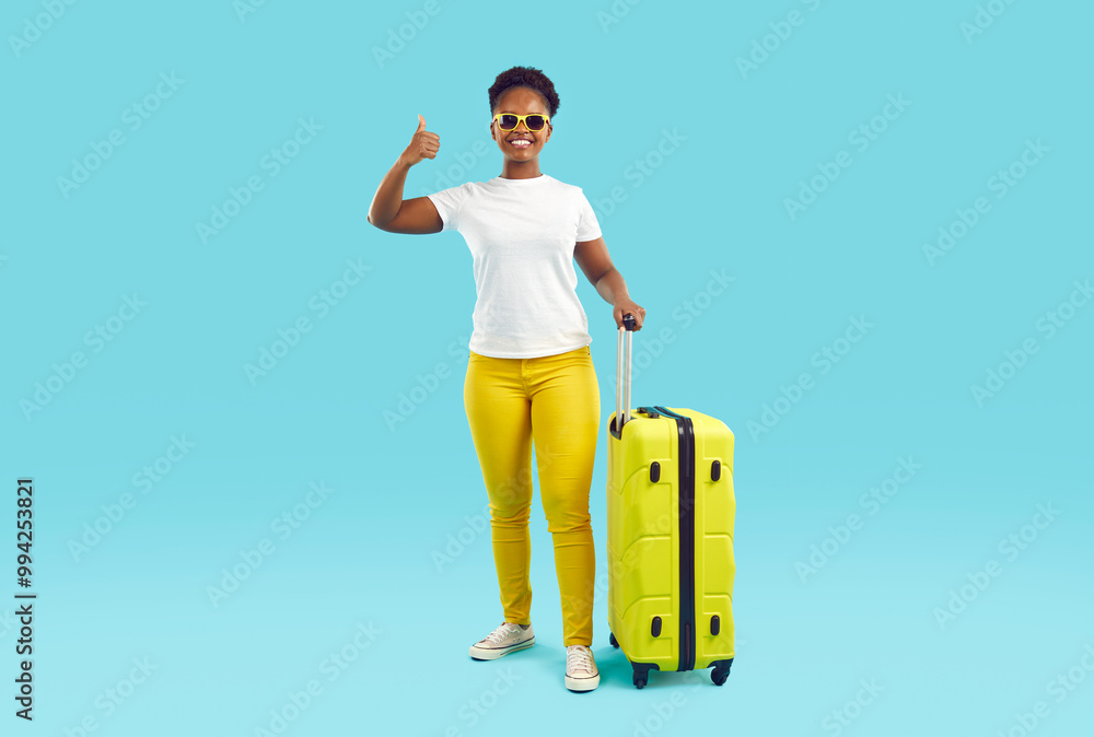 Full length portrait of joyful smiling african american woman in yellow pants and white t-shirt showing thumb up with yellow suitcase on turquoise background. Travelling, tourism journey trip concept Full length portrait of joyful smiling african american woman in yellow pants and white t-shirt showing thumb up with yellow suitcase on turquoise background. Travelling, tourism journey trip concept