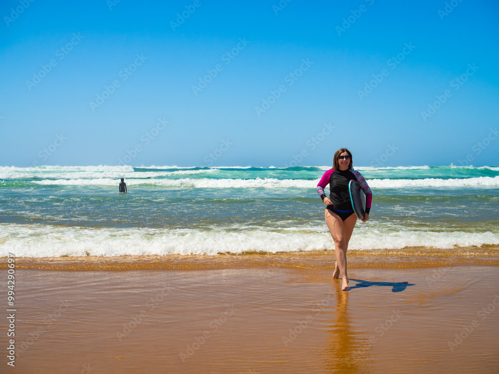 Water sports. Beautiful long-haired woman wearing long sleeve swimsuit holding multicolored bodyboard running out of water on sunny sandy beach. Front view. Bordeira praia, Algarve, Portugal. Water sports. Beautiful long-haired woman wearing long sleeve swimsuit holding multicolored bodyboard running out of water on sunny sandy beach. Front view. Bordeira praia, Algarve, Portugal.