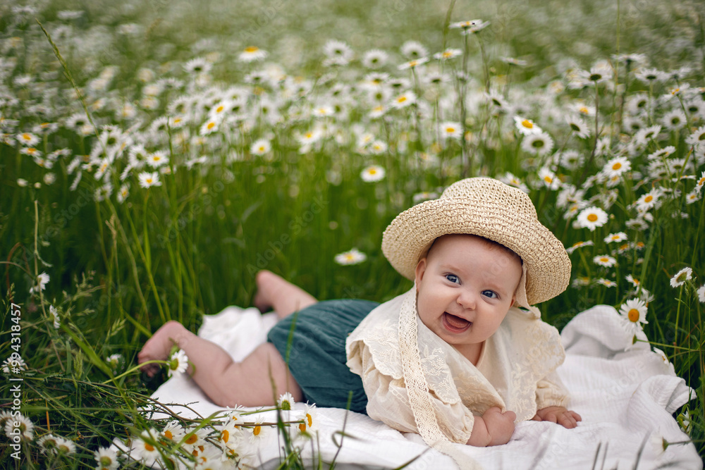 beautiful chubby little girl infant baby lying in a field with daisies in a hat on a summer afternoon in nature