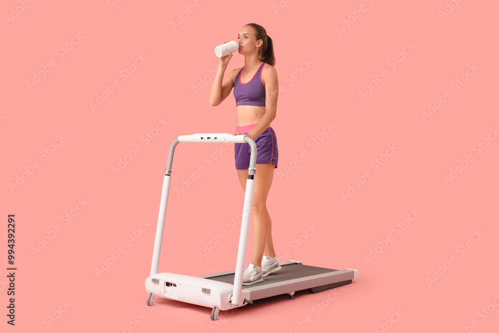 Sporty young woman drinking water on treadmill against pink background Sporty young woman drinking water on treadmill against pink background