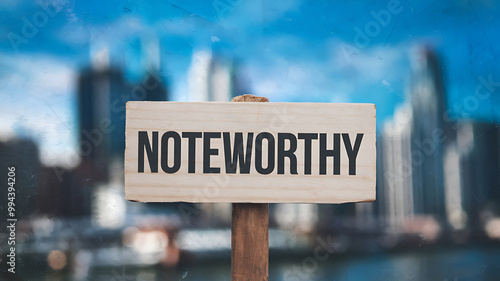 A wooden sign with black text saying NOTEWORTHY against a blurred city skyline background, conveying a message of importance and achievement. 
