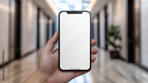 A hand presents a blank smartphone against a sleek corridor, highlighting the device's simple design. The surrounding environment features polished floors and contemporary architecture