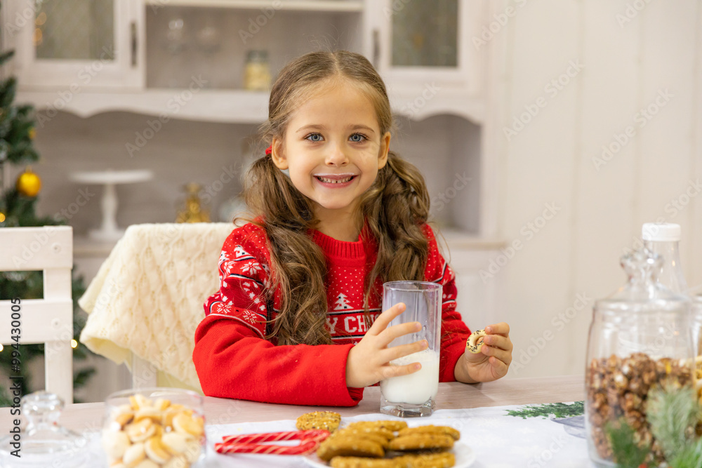 Happy joyful cute little girl in red sweater drinking milk and eating delicious cookie near Christmas tree in the kitchen for Christmas morning. Christmas time. Christmas holidays Happy joyful cute little girl in red sweater drinking milk and eating delicious cookie near Christmas tree in the kitchen for Christmas morning. Christmas time. Christmas holidays