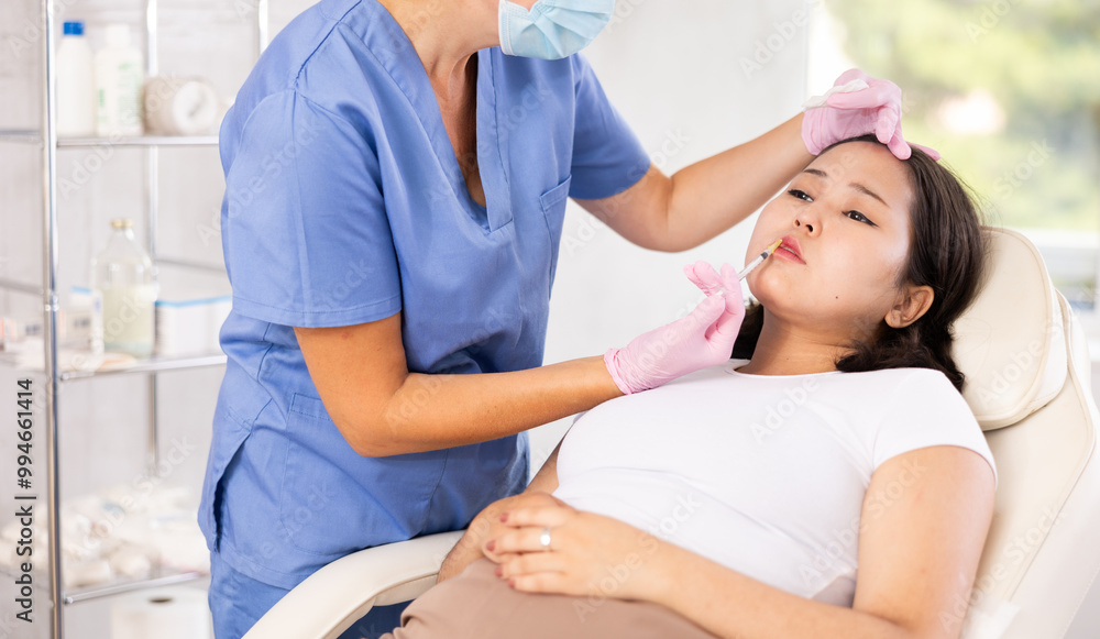Fototapeta premium Closeup face of young female client receiving injections during lip enhancement procedure, professional cosmetologist hands in rubber gloves holding syringe