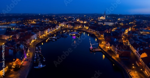 An aerial view of a city at night, with the lights of the buildings reflecting in the water.