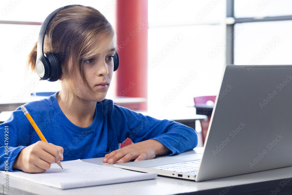 In school, boy wearing headphones writing in notebook while using laptop In school, boy wearing headphones writing in notebook while using laptop