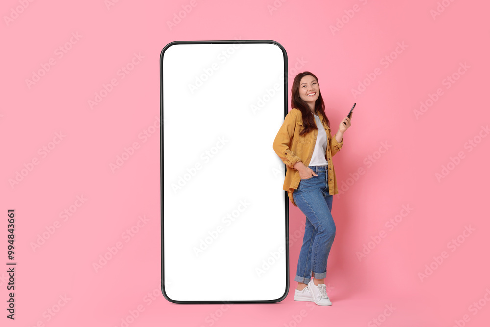 Happy smiling woman with smartphone standing near big mobile phone with blank screen on pink background. Mockup for design Happy smiling woman with smartphone standing near big mobile phone with blank screen on pink background. Mockup for design