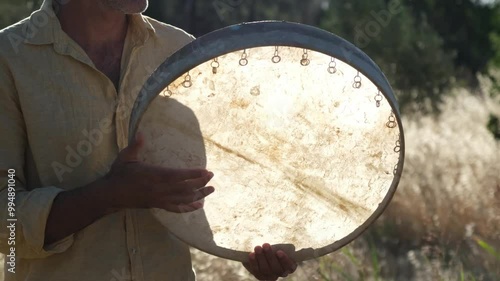 A Middle Eastern man in his forties is seen playing the bendir in an olive field. The traditional music blends with the natural setting, capturing the peaceful atmosphere photo