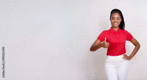 A portrait of a Black woman giving a thumbs up while wearing a red polo shirt and white skinny pants against a background of white walls.