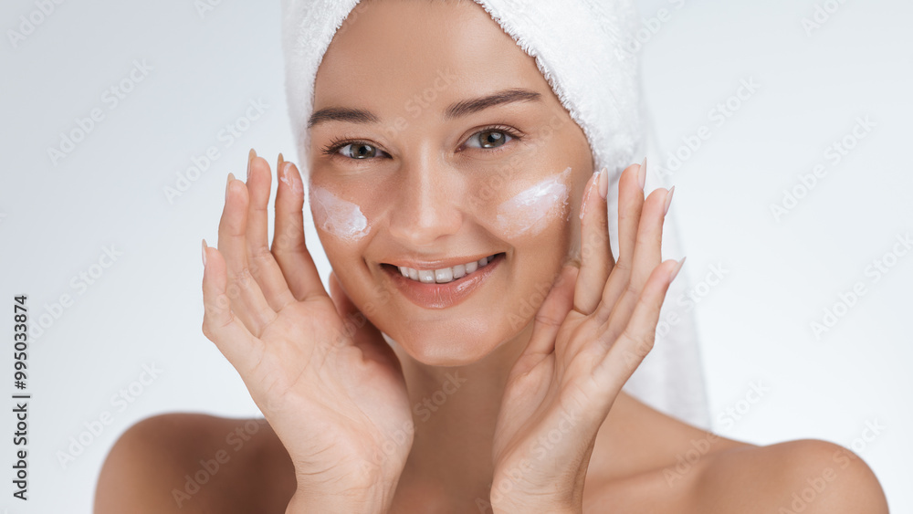 Smiling Woman with Towel Wrapped Head Applying Skincare Cream to Cheeks Smiling Woman with Towel Wrapped Head Applying Skincare Cream to Cheeks