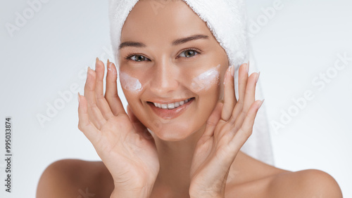 Smiling Woman with Towel Wrapped Head Applying Skincare Cream to Cheeks