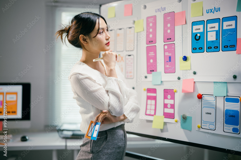 Young professional is analyzing the layout of a mobile application, holding a pen in her hand and some color samples in the other, while standing in her office Young professional is analyzing the layout of a mobile application, holding a pen in her hand and some color samples in the other, while standing in her office