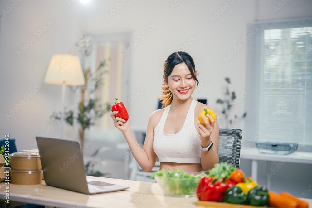 Young woman is smiling while choosing between a red and a yellow bell pepper in her kitchen, with a laptop and a bowl of fresh vegetables on the table Young woman is smiling while choosing between a red and a yellow bell pepper in her kitchen, with a laptop and a bowl of fresh vegetables on the table
