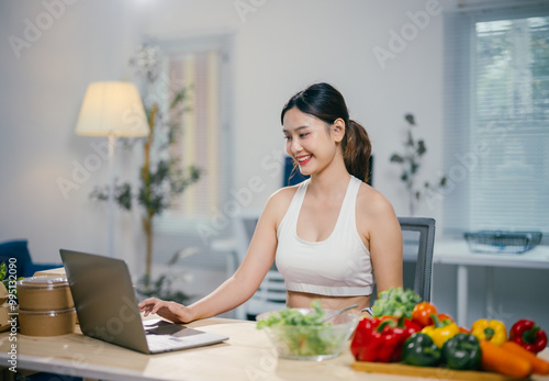 Young woman in sportswear using laptop while having fresh vegetables salad in the kitchen at home, healthy food and technology concept