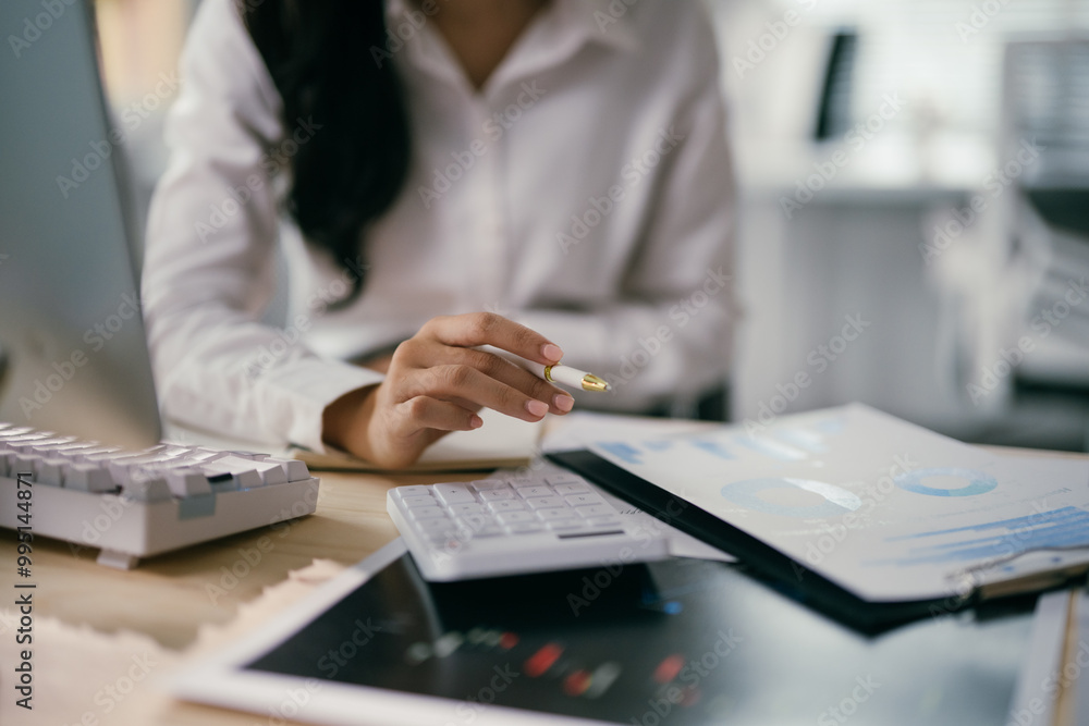 Fototapeta premium Focused businesswoman reviewing financial data on computer at office desk, surrounded by technology and paperwork, strategizing for success