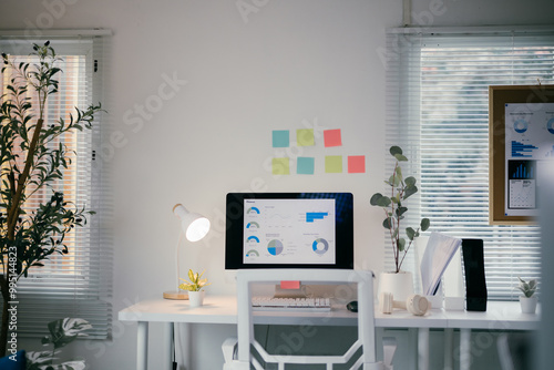 Modern, bright home office is pictured with a computer monitor displaying analytics charts and graphs. The desk is white with a white chair, keyboard, mouse, lamp, and potted plants