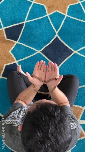 Top view photo of a Muslim man praying while holding prayer beads and raising both hands in a mosque