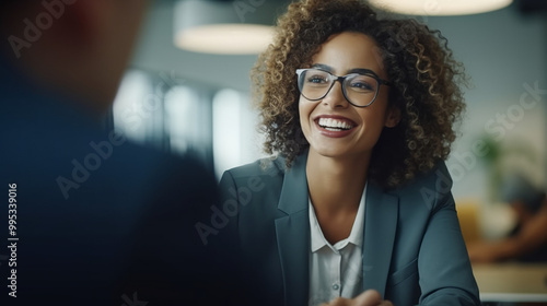 photograph of a smiling female executive with curly hair and glasses, wearing business attire, having an interview in the office room, while two people sit opposite her. 