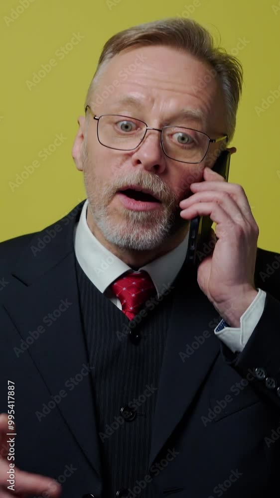Nervous middle aged businessman with a cellphone. Mature man in dark suit talking on the phone and giving instructions to somebody on yellow background. Vertical video