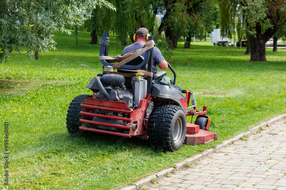 A person operating a lawn mower in a park, cutting the grass. The person is wearing a hat and headphones, focused on the task at hand A person operating a lawn mower in a park, cutting the grass. The person is wearing a hat and headphones, focused on the task at hand