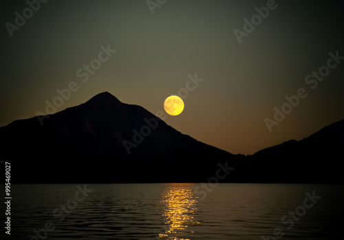 Red moon reflecting on a serene mountain lake at night, with distant mountains on the horizon under the dark sky.