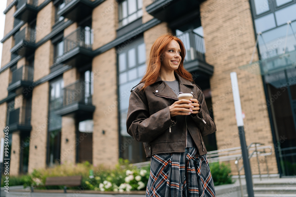 Low-angle view of happy redhead woman savoring coffee break in urban environment, holding cup and sipping drink with smile, exuding joy and relaxation, standing on background of modern building. Low-angle view of happy redhead woman savoring coffee break in urban environment, holding cup and sipping drink with smile, exuding joy and relaxation, standing on background of modern building.