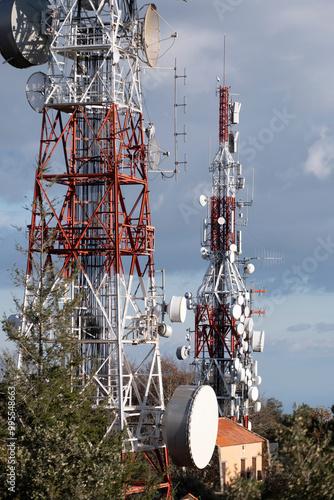 Two tall transmission towers with multiple antennas and satellite dishes stand amidst a natural environment, under a partly cloudy sky. photo