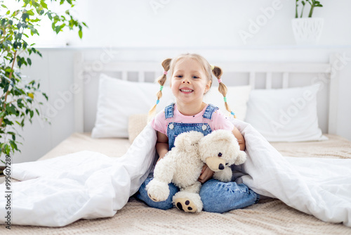 a small child, a blonde girl, holds a teddy bear in her hands under a blanket and smiles on a white bed in a bright bedroom