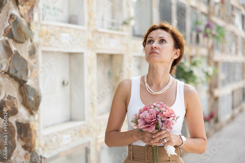 Elegant grieving woman in forties, wearing white tank top and pearls, standing with bouquet in hands near stone columbarium wall in cemetery, gazing sorrowfully and contemplating loss photo