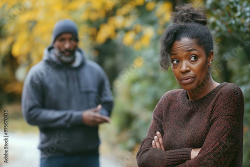 A woman stands with crossed arms in a park, facing a man as autumn leaves create a colorful backdrop during a tense conversation photo