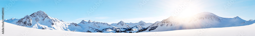 A wide panoramic shot of a snow-covered mountain range under a bright, clear sky. The jagged mountain peaks are blanketed in thick snow, their edges sharply defined against the vibrant backdrop. The A wide panoramic shot of a snow-covered mountain range under a bright, clear sky. The jagged mountain peaks are blanketed in thick snow, their edges sharply defined against the vibrant backdrop. The