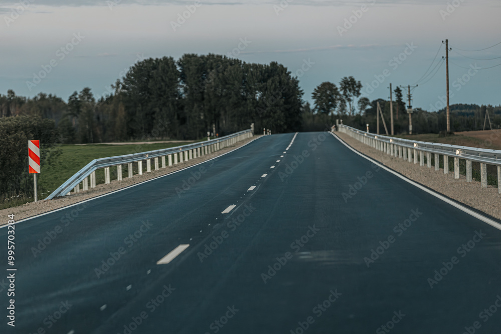Fototapeta premium A smooth, empty road winds through a wooded area with dense trees on one side and open fields on the other. The soft evening light creates a peaceful atmosphere with ample copy space in the sky and on