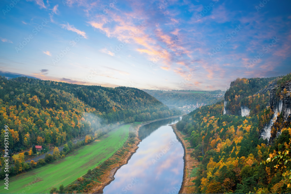 Fototapeta premium Scenic view of the Elbe River winding through colorful autumn forests in Saxon Switzerland National Park, Germany. Misty valley under a dramatic sunrise sky