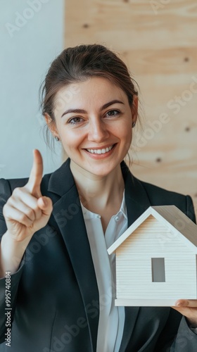 A female real estate agent in business attire with hair tied back, smiling confidently while pointing at a white model house in a light-colored office setting