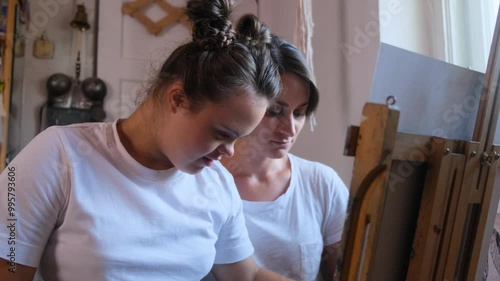 A young woman with Down syndrome is looking intently at her mother, who is explaining something to her. The two are in an art studio, with a wooden easel in the background.