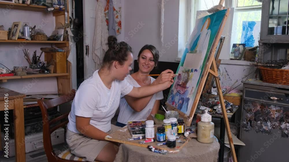 Two young women are painting together in an art studio. One woman has Down syndrome, and she is holding a paintbrush and looking at the canvas. Two young women are painting together in an art studio. One woman has Down syndrome, and she is holding a paintbrush and looking at the canvas.