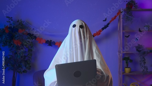 Photography Funny halloween ghost in white sheet with black eyes in dark room working on laptop, typing text surfing on the web