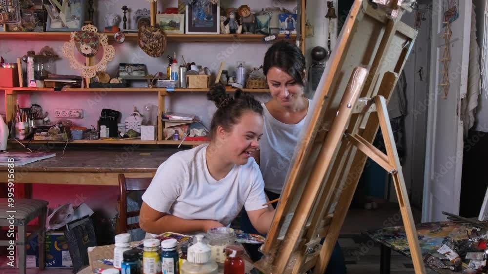 A young woman with Down syndrome is painting on a canvas, guided by her friend. They are both focused on their artistic project, with the woman with Down syndrome looking intently at the canvas. A young woman with Down syndrome is painting on a canvas, guided by her friend. They are both focused on their artistic project, with the woman with Down syndrome looking intently at the canvas.