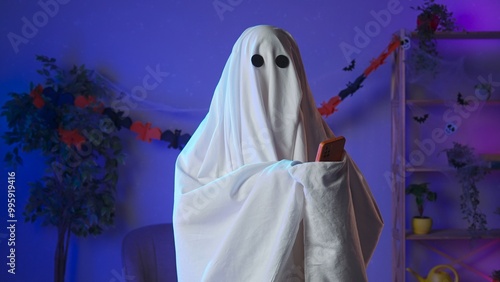 Canvastavla Funny halloween ghost in white sheet with black eyes in dark room talking on smartphone by video call, positive expression