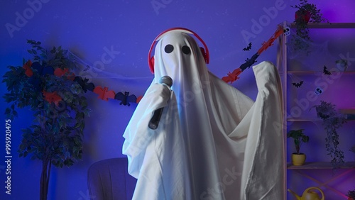 Funny halloween ghost in white sheet with black eyes in dark room dancing to music in headphones sings in karaoke microphone. Happy halloween spooky concept. photo