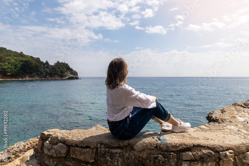Young woman in blue jeans and white blouse sitting on rock ledge looking out to ocean with scenic rocky coastline in background. Copy space.travel or lifestyle content