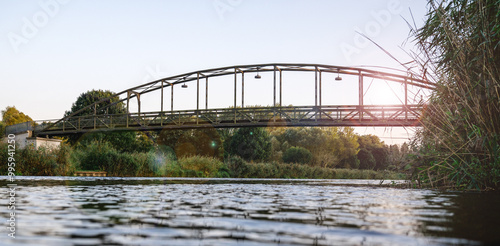 eine stählerne Fußgängerbrücke, die sich über einen ruhigen Fluss oder Kanal spannt, umgeben von üppigem Grün, mit Sonnenlicht, das eine friedliche Atmosphäre schafft photo