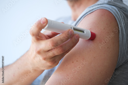an anonymous adult man injecting himself a treatment such as insulin or Adalimumab, Etanercept or epinephrine into his shoulder. It is a white pen injector with red head for health care