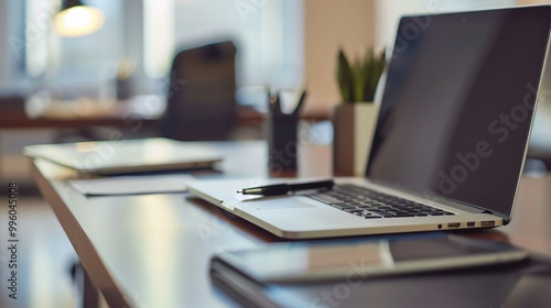 A laptop on a desk in an office with a pen, a plant, and another laptop in the background.