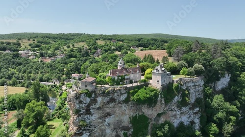 A breathtaking aerial view of a historic village nestled in lush green hills. The scene features a beautiful castle  on a cliffside, overlooking a river flowing through the valley. 