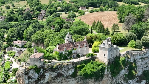 A breathtaking aerial view of a historic village nestled in lush green hills. The scene features a beautiful castle  on a cliffside, overlooking a river flowing through the valley. 