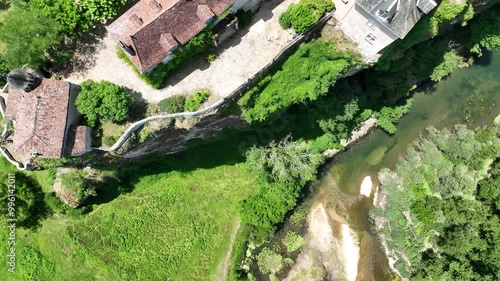 A breathtaking aerial view of a historic village nestled in lush green hills. The scene features a beautiful castle  on a cliffside, overlooking a river flowing through the valley. 