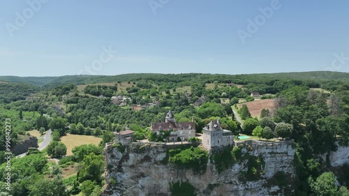A breathtaking aerial view of a historic village nestled in lush green hills. The scene features a beautiful castle  on a cliffside, overlooking a river flowing through the valley. 