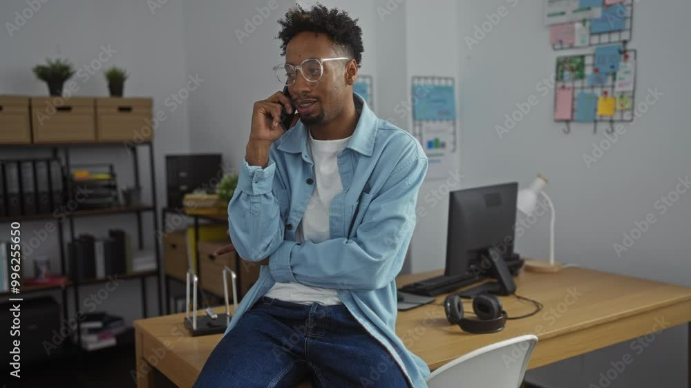 A handsome black man with a beard is talking on the phone in an indoor office setting, surrounded by work-related items and decor. A handsome black man with a beard is talking on the phone in an indoor office setting, surrounded by work-related items and decor.