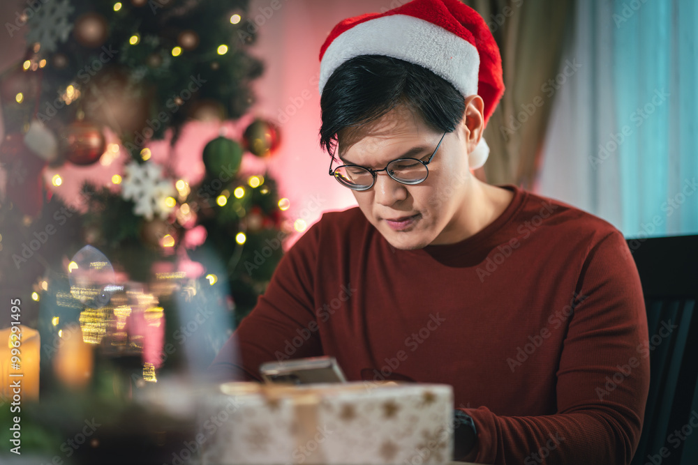 Asian man in a Santa hat enjoying a quiet Christmas evening, texting on his smartphone, surrounded by festive holiday lights, wine, and gifts, capturing the warmth of holiday celebrations at home. Asian man in a Santa hat enjoying a quiet Christmas evening, texting on his smartphone, surrounded by festive holiday lights, wine, and gifts, capturing the warmth of holiday celebrations at home.